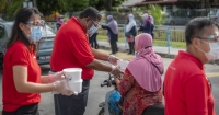 A medium full landscape shot of Sunwayians in signature red polo tee and face masks handing over containers of #SunwayforGood bubur lambuk to commuters during Sunway Group’s Raya Cheer 2021