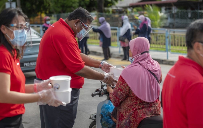 A medium full landscape shot of Sunwayians in signature red polo tee and face masks handing over containers of #SunwayforGood bubur lambuk to commuters during Sunway Group’s Raya Cheer 2021