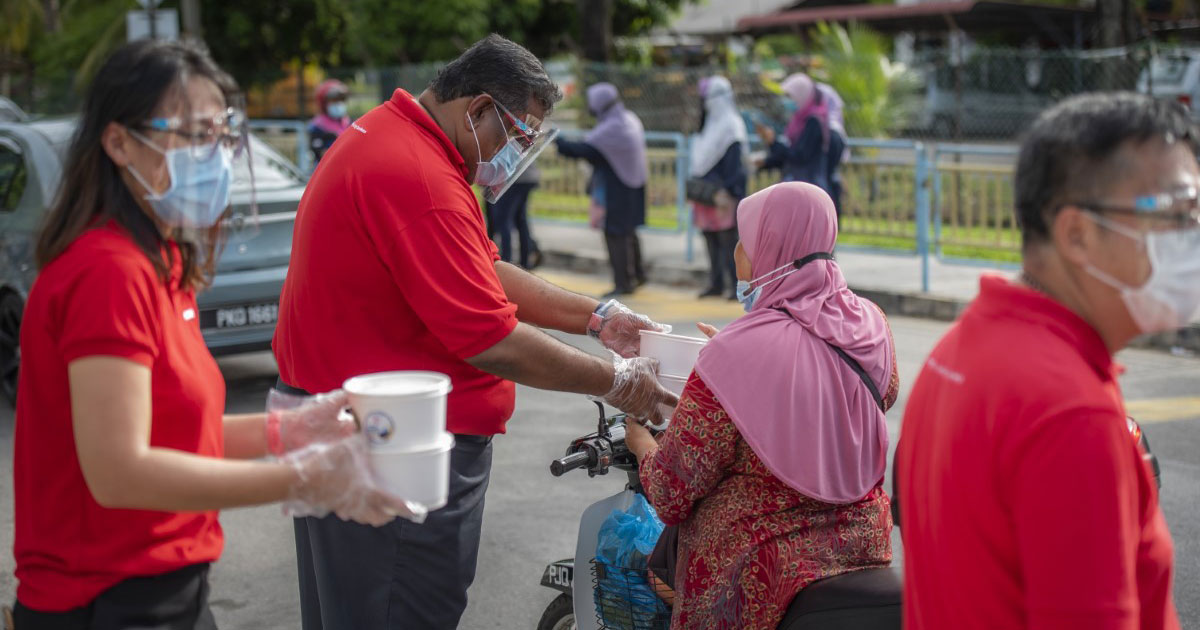 A medium full landscape shot of Sunwayians in signature red polo tee and face masks handing over containers of #SunwayforGood bubur lambuk to commuters during Sunway Group’s Raya Cheer 2021