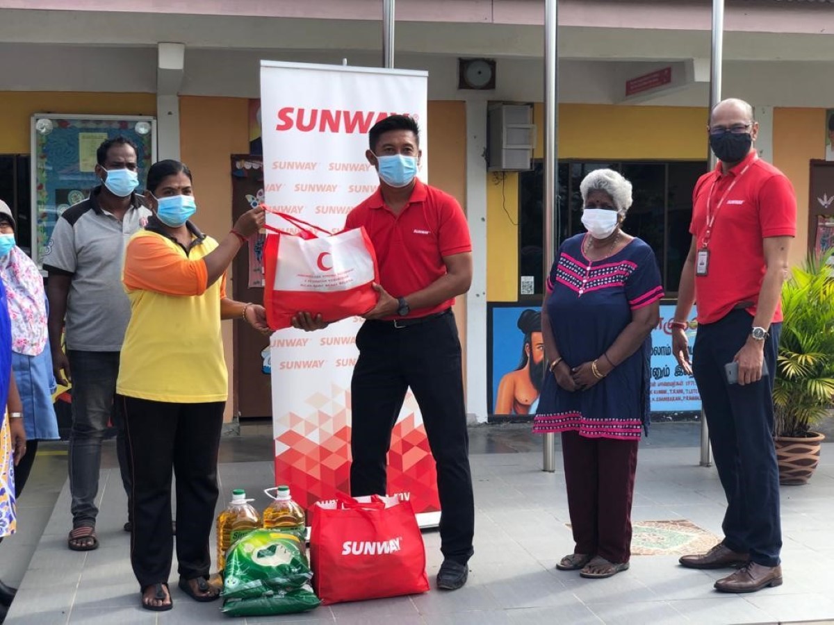A full landscape shot of Sunway Lost World of Tambun general manager Nurul Nuzairi bin Mohd Azahari in signature red polo tee and face mask handing over bags of goodies to beneficiaries, as Sunway Group general manager of CSR, Internal Branding and Events, Bernard Paul looks ahead under broad daylight