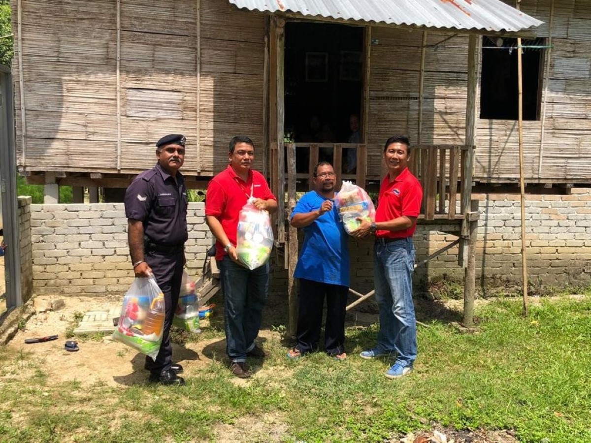 A full landscape shot of Sunway Lost World of Tambun general manager Nurul Nuzairi bin Mohd Azahari in signature red polo tee and vibrant smile handing bags of goodies to villagers outside a traditional village house under bright sunlight at Kampung Orang Asli Sungai Choh