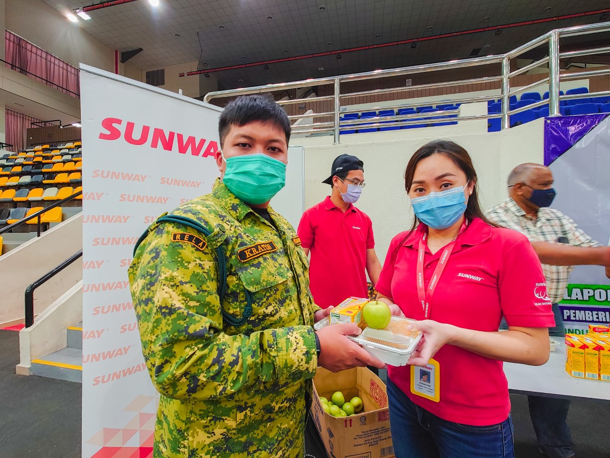 Dressed in signature red Sunway polo, lanyard and face mask, Susan Sae of Sunway Property Ipoh sales and marketing department hands over a box of nutritious meal and a bright green apple to a RELA officer in face mask at the Stadium Indera Mulia PPV in Ipoh