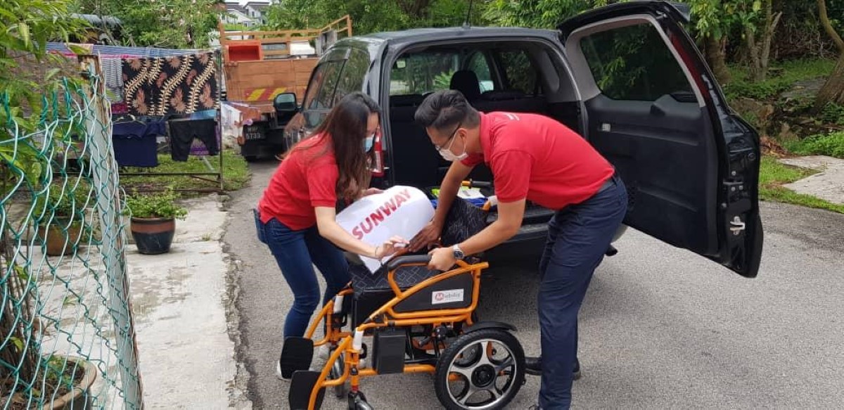 A full landscape shot of Susan (left) in signature red polo tee and face mask holding a #SunwayforGood banner while unloading wheelchairs outside a household compound in the northern region