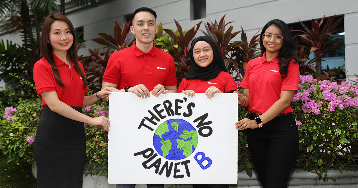 Empat kakitangan Sunway - seorang lelaki, dan tiga perempuan - memegang poster yang menyatakan "Tiada Planet B" dan gambar dunia, di tengah-tengah latar belakang hijau yang subur.