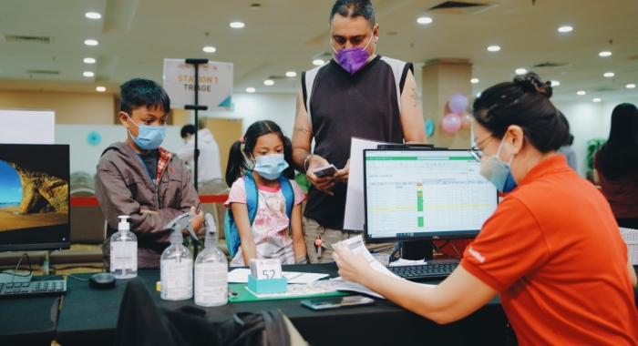 A medium full landscape shot of a Sunwayian in signature red Sunway polo and face mask behind a lit computer screen, attending to two children and a parent up front at Sunway Medical Centre, with bottles of hand sanitisers and various tools and paperwork on the table between them