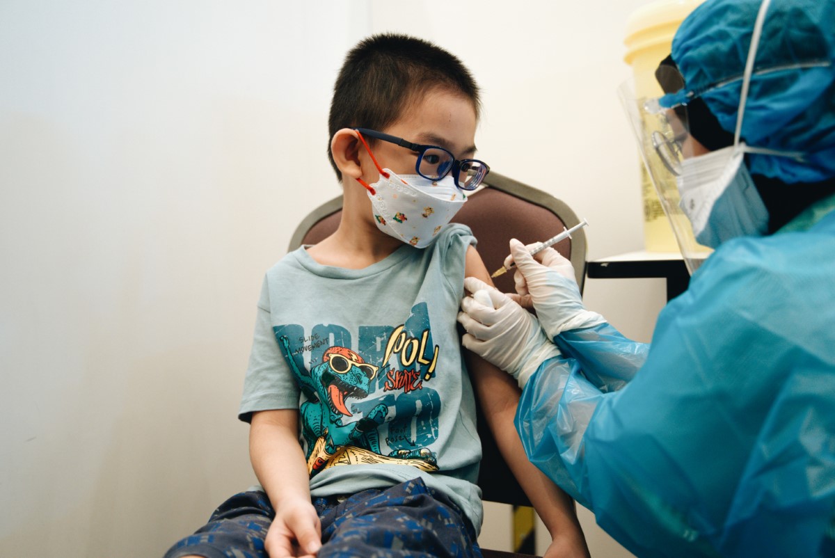 A medium landscape shot of a child in glasses boldly receiving his vaccination injection administered by a Sunway Medical Centre personnel dressed in complete protective gear