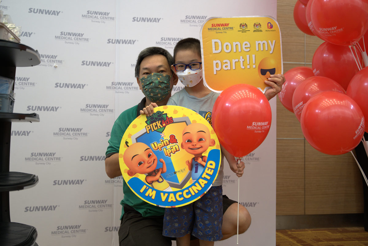A medium full landscape shot of a boy posing for a commemoratory shot with his father, holding balloons and statement props after his first COVID-19 vaccination at Sunway Medical Centre