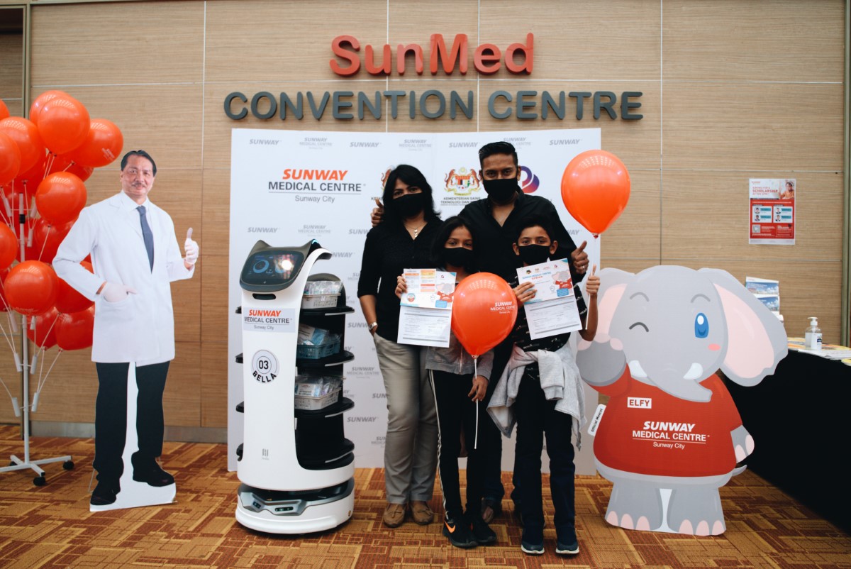 A full landscape shot of a family of four in black and grey posing alongside BellaBot, a cutout of Elfy as well as a cutout of Tan Sri Dato' Seri Dr. Noor Hisham Abdullah, among red balloons and banners, where the children in front held up their respective certificates of vaccination at Sunway Medical Centre