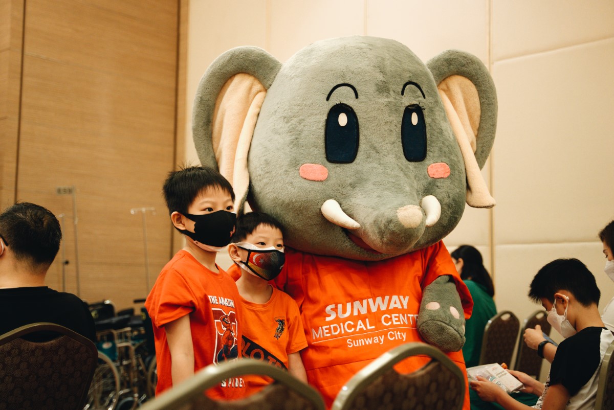 A medium landscape shot of two children in red posing for a picture with Sunway Medical Centre’s mascot Elfy, amidst a crowd practicing social distancing in a hall