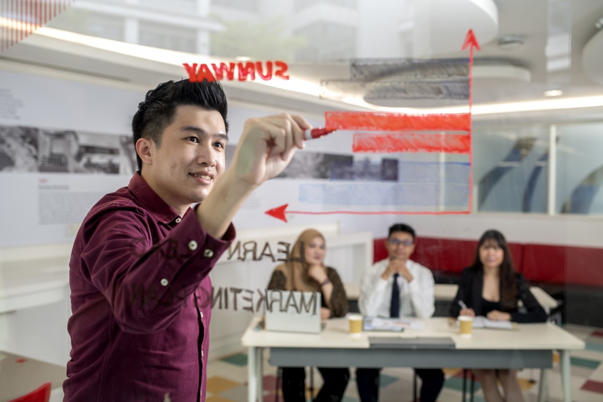 A side-angle shot of a Sunway employee mapping out a marketing graph with three other employees watching him at the background