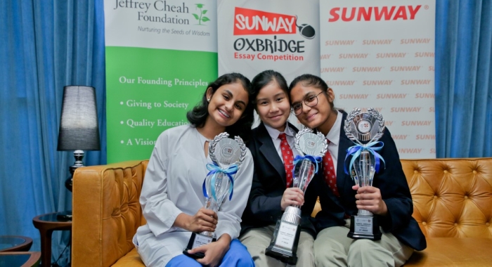 A medium full landscape shot of A full landscape shot of Ariana Carmel Ravi, Arianna Saiful and Harvynna Kaur Kler seated side-by-side on a plush tan couch, holding up award trophies in front of Jeffrey Cheah Foundation, Sunway-Oxbridge Essay Competition and Sunway Group’s standing banners