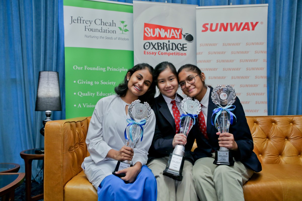 A medium full landscape shot of A full landscape shot of Ariana Carmel Ravi, Arianna Saiful and Harvynna Kaur Kler seated side-by-side on a plush tan couch, holding up award trophies in front of Jeffrey Cheah Foundation, Sunway-Oxbridge Essay Competition and Sunway Group’s standing banners