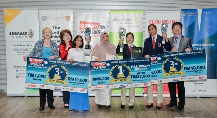 A full landscape shot of Ariana Carmel Ravi, Arianna Saiful and Harvynna Kaur Kler holding up award trophies and mock cheques on-stage at the Sunway-Oxbridge Essay Competition prize-giving ceremony in July 2019, alongside executive committee member and past president of the Oxford & Cambridge Society Malaysia, Margaret Hall, Sunway Education Group chief executive officer Dr. Elizabeth Lee and Ministry of Education’s English Language Teaching Centre director Yang Berusaha Farah Mardhy binti Aman
