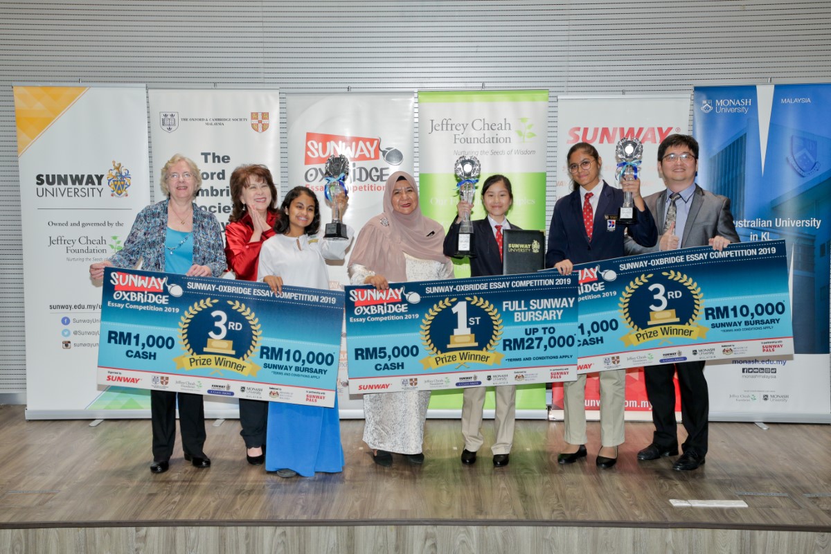 A full landscape shot of Ariana Carmel Ravi, Arianna Saiful and Harvynna Kaur Kler holding up award trophies and mock cheques on-stage at the Sunway-Oxbridge Essay Competition prize-giving ceremony in July 2019, alongside executive committee member and past president of the Oxford & Cambridge Society Malaysia, Margaret Hall, Sunway Education Group chief executive officer Dr. Elizabeth Lee and Ministry of Education’s English Language Teaching Centre director Yang Berusaha Farah Mardhy binti Aman