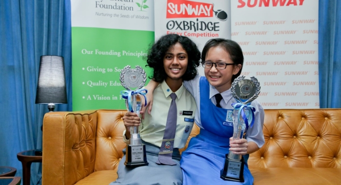 A medium full landscape shot of A full landscape shot of Asfa Nisha binti Basheer Ali and Abel (previously Avril) Yong Zhi Ying seated side-by-side on a plush tan couch, holding up award trophies in front of Jeffrey Cheah Foundation, Sunway-Oxbridge Essay Competition and Sunway Group’s standing banners