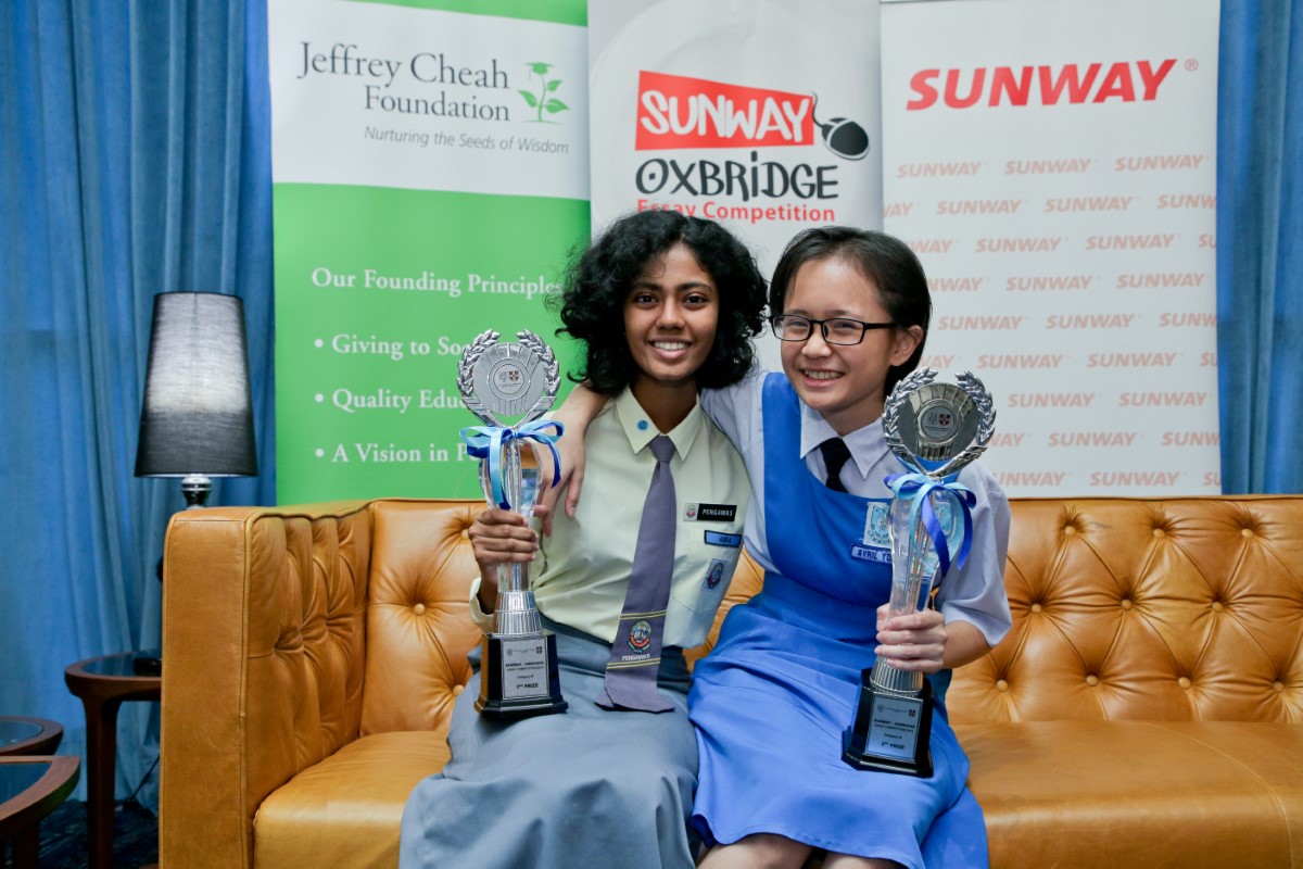 A medium full landscape shot of A full landscape shot of Asfa Nisha binti Basheer Ali and Abel (previously Avril) Yong Zhi Ying seated side-by-side on a plush tan couch, holding up award trophies in front of Jeffrey Cheah Foundation, Sunway-Oxbridge Essay Competition and Sunway Group’s standing banners
