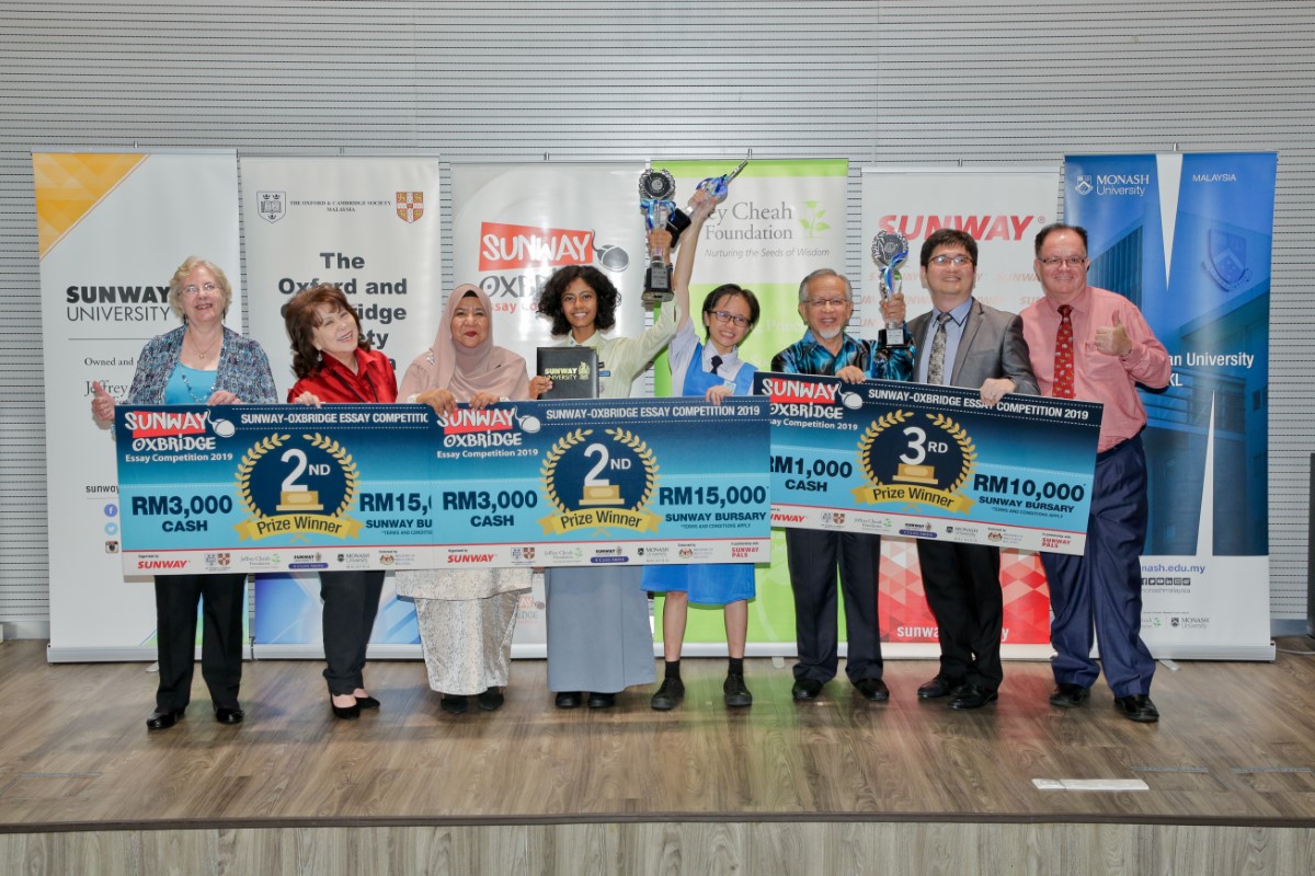 A full landscape shot of Asfa Nisha binti Basheer Ali, Abel (previously Avril) Yong Zhi Ying and Muhammad Luqman Mohammad Nawar’s grandfather holding up award trophies and mock cheques on-stage at the Sunway-Oxbridge Essay Competition prize-giving ceremony in July 2019, alongside executive committee member and past president of the Oxford & Cambridge Society Malaysia, Margaret Hall, Sunway Education Group chief executive officer Dr. Elizabeth Lee and Ministry of Education’s English Language Teaching Centre director Yang Berusaha Farah Mardhy binti Aman
