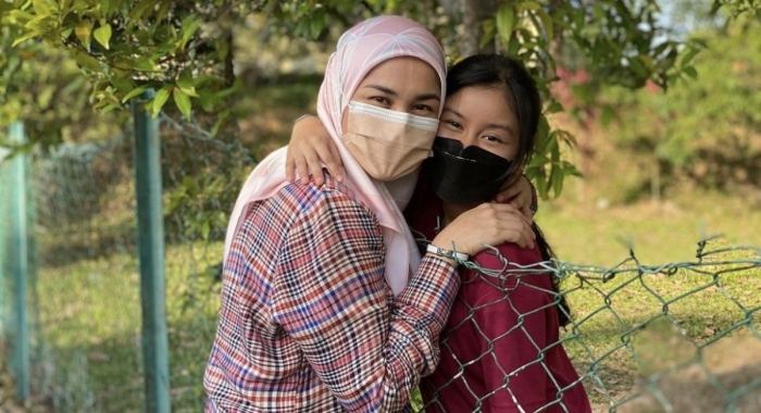 A medium landscape shot of Arianna Saiful hugging her mother over a wired fence, surrounded by trees and sunlight