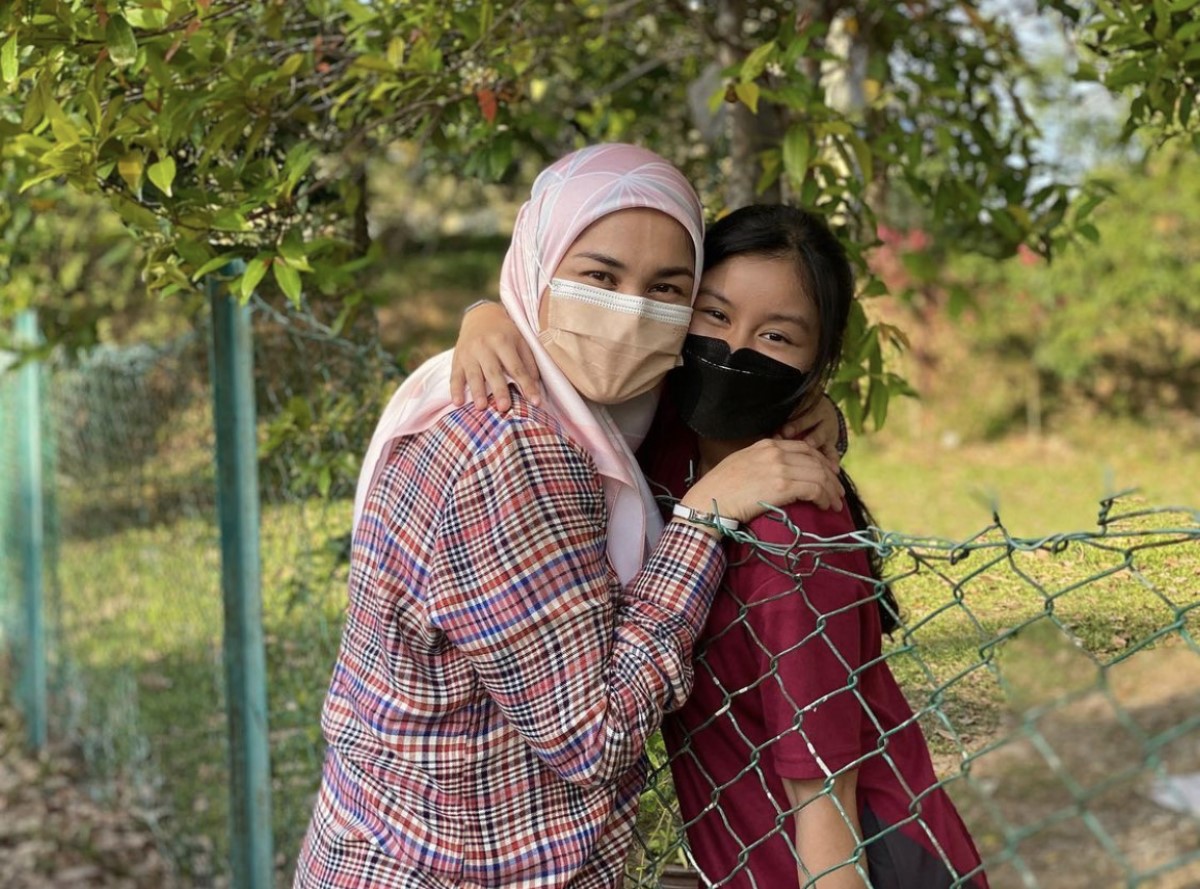 A medium landscape shot of Arianna Saiful hugging her mother over a wired fence, surrounded by trees and sunlight