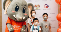 A medium full landscape shot of three children in masks posing with Sunway Medical Centre’s mascot, Elfy, amidst huge banners and red balloons in preparation for their vaccination under Malaysia’s PICKids