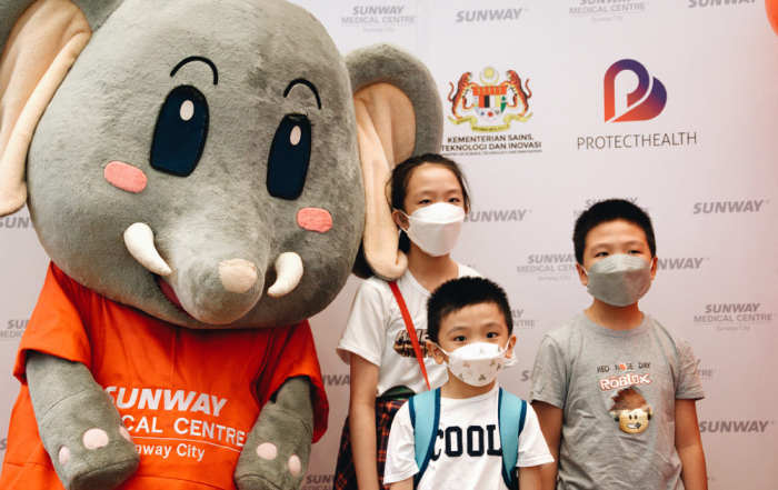 A medium full landscape shot of three children in masks posing with Sunway Medical Centre’s mascot, Elfy, amidst huge banners and red balloons in preparation for their vaccination under Malaysia’s PICKids