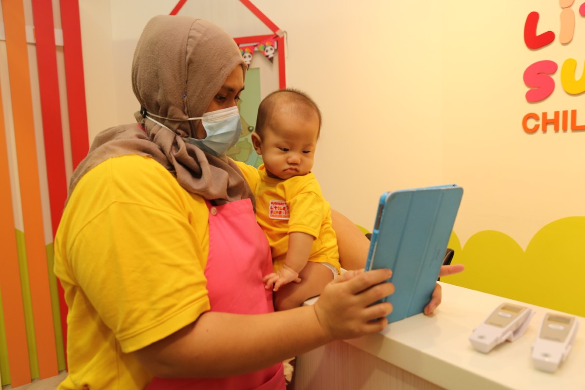 A tight medium landscape shot of a Sunway Little Sunshine nursery caregiver helping an infant check-in the Oodlins app upon arrival