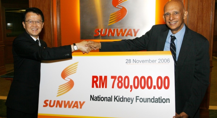 Tan Sri Sir Jeffrey Cheah handing a mock cheque with the second logo to the National Kidney Foundation, and shaking hands with the Sunway logo in the background