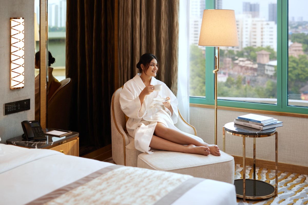 A female in a bathrobe enjoying a cup of coffee, lounging on a comfortable sofa with bed and books beside her. The female is looking out to the view of Sunway City Kuala Lumpur.