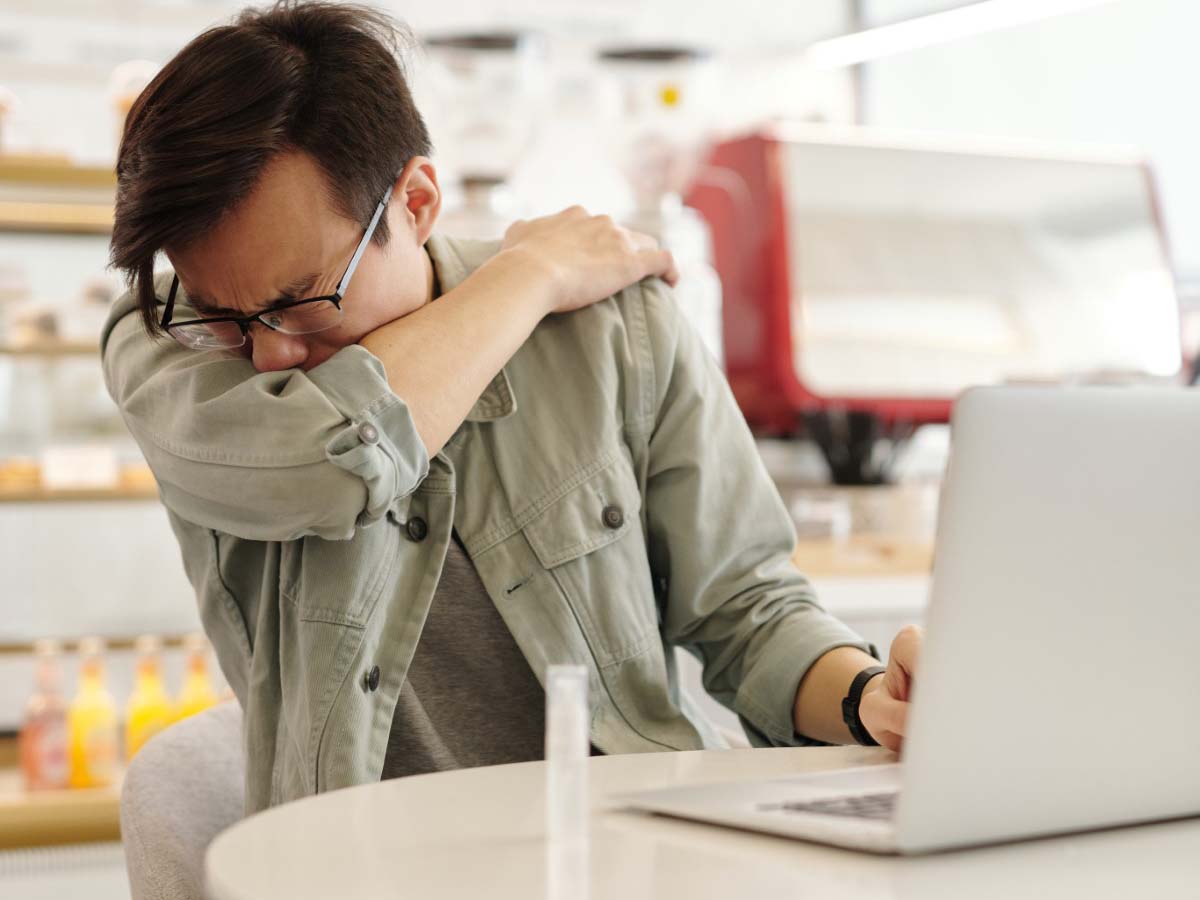 A man in a grey jacket sneezing, with a laptop in front of him