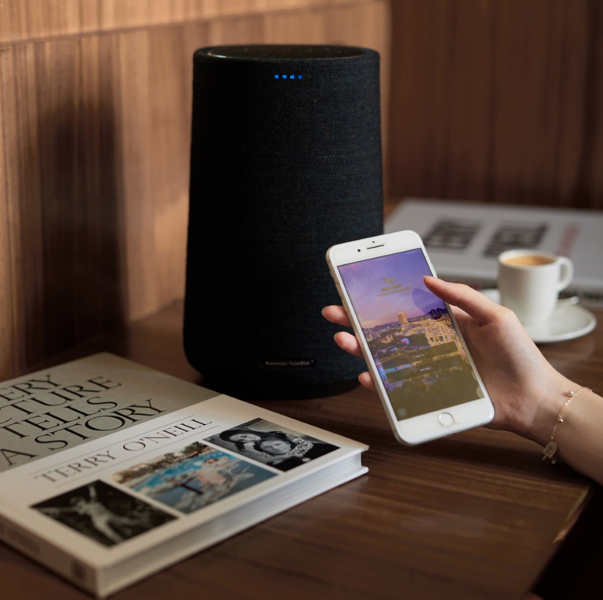 A hand holding a smartphone, connecting to the Harman Kardon speakers. Alongside it is a book and cup and saucer.