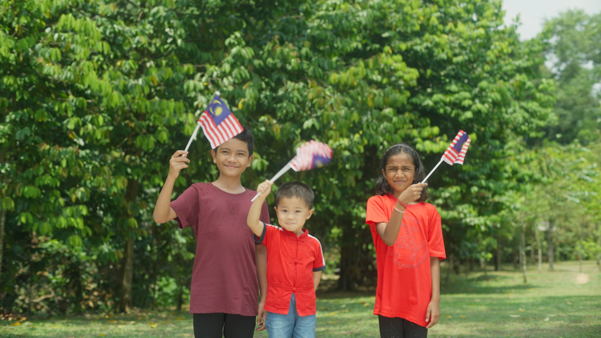 Three children – an Indian boy, a Chinese girl and a Malay girl – waving Malaysian flags, with trees in the background.