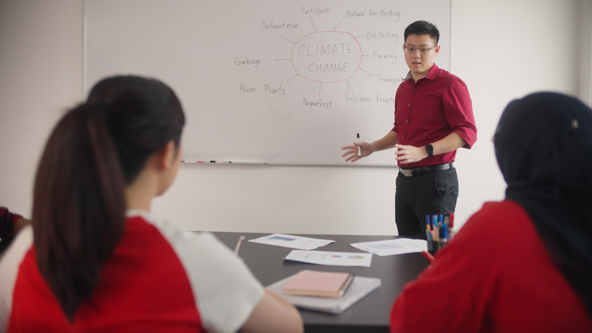 A male lecturer is mentoring and teaching the Junior Rangers, drawing items on the white board. Notes and books are spread across the table in front as well.