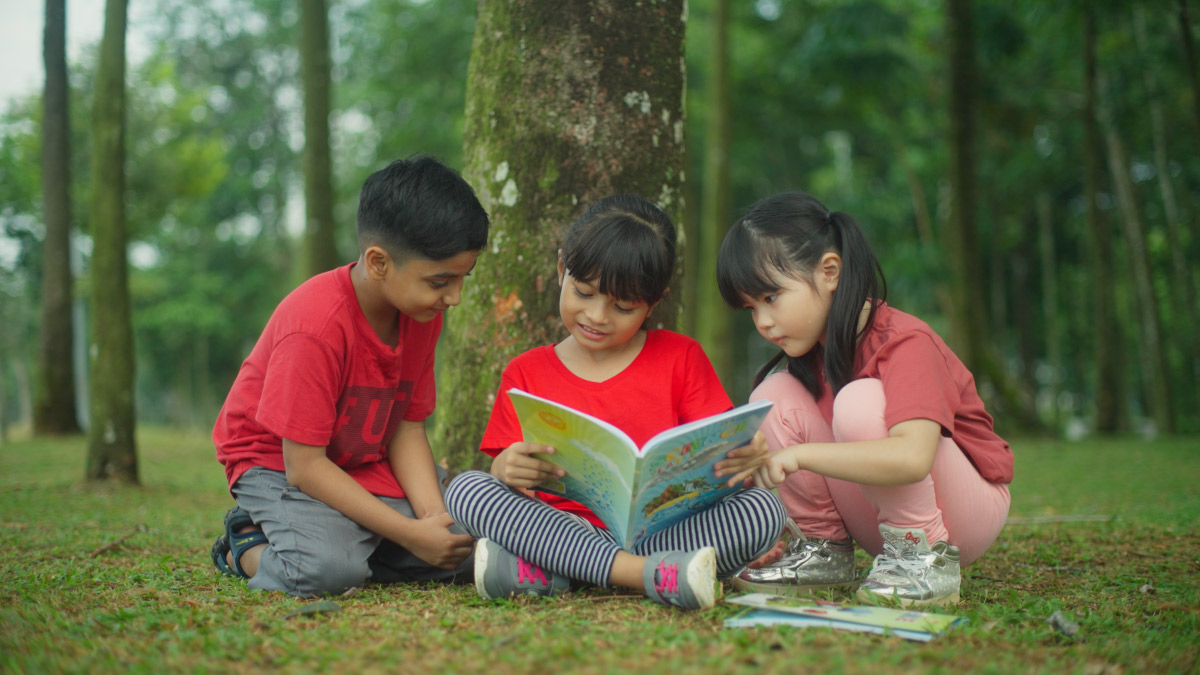 Three children – an Indian boy, a Chinese girl and a Malay girl – shares and reads a book by a tree.