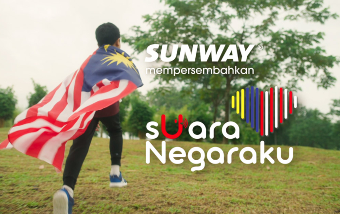 A boy running up a hill with the Malaysian flag draped across his back, with trees in the background. Also features the Sunway logo, ”Suara Negaraku” tagline and the heart-shaped Malaysia-themed key visual.