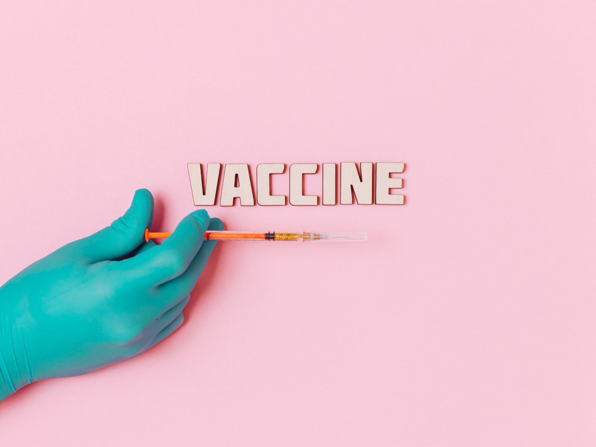 A pastel pink background, with a gloved hand with an injection syringe and the words ”Vaccine” displayed there