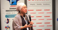 Prof. Tan Sri Dr. Jemilah holding a microphone and a clicker, giving a presentation amidst the backdrop of Sunway University and DHL bunting