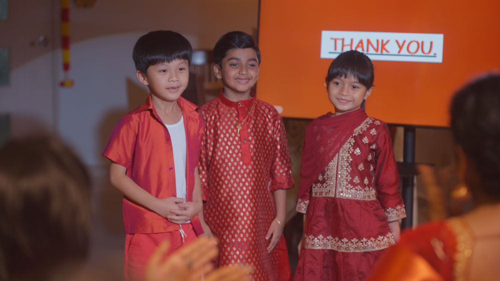 Kishen and his friends are giving a presentation to their families. Clothed in red Deepavali traditional clothing and the spotlight on them during their presentation.