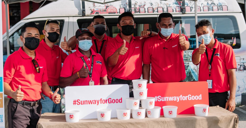 A medium landscape shot of Sunway volunteers in their classic red polo tee, flashing thumbs up behind #SunwayforGood signs propped up against bubur lambuk containers during Raya Cheer 2022.