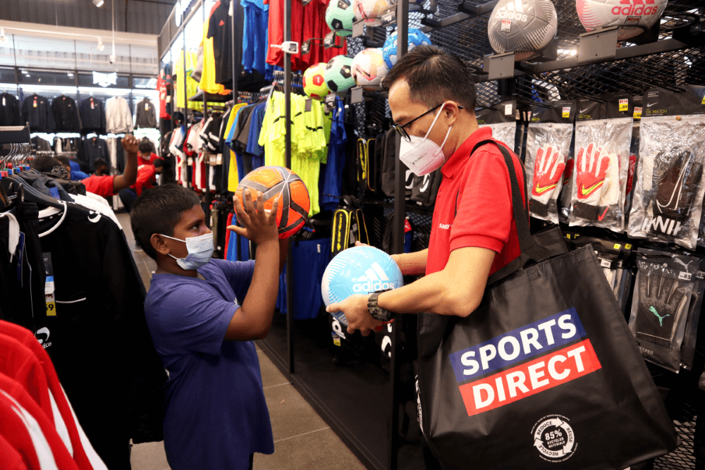 A sunway staff wearing glasses and a young boy at Sports Direct store, surveying footballs of different colours such as light blue or orange.