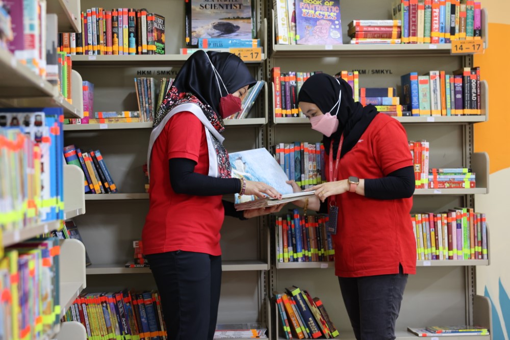 A medium full landscape shot of two Sunway volunteers in classic red polo, leafing through pages of donated books in a newly-furnished library at Sekolah Menengah Kebangsaan Yaacob Latif in Cheras, Kuala Lumpur.