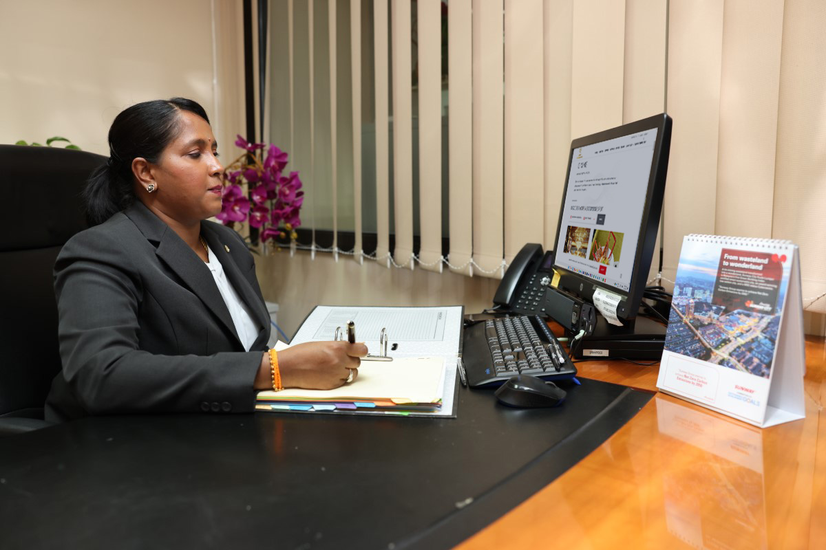 Shanta at work. At her desk, she is writing with a pen and a monitor in front of her.