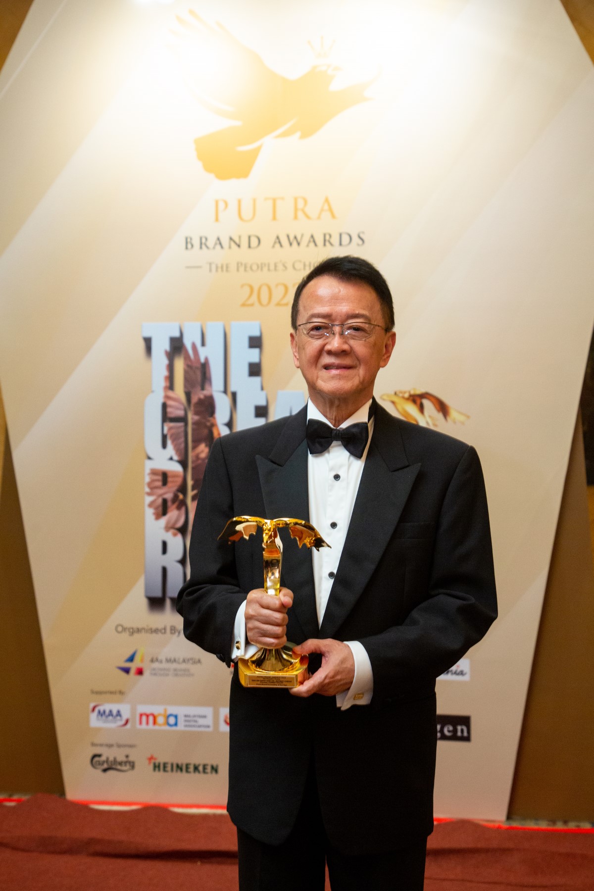 Tan Sri Dr. Jeffrey Cheah holding up the Putra Brand Awards trophy – shaped in the golden eagle amidst the gold Putra Brand Awards backdrop.