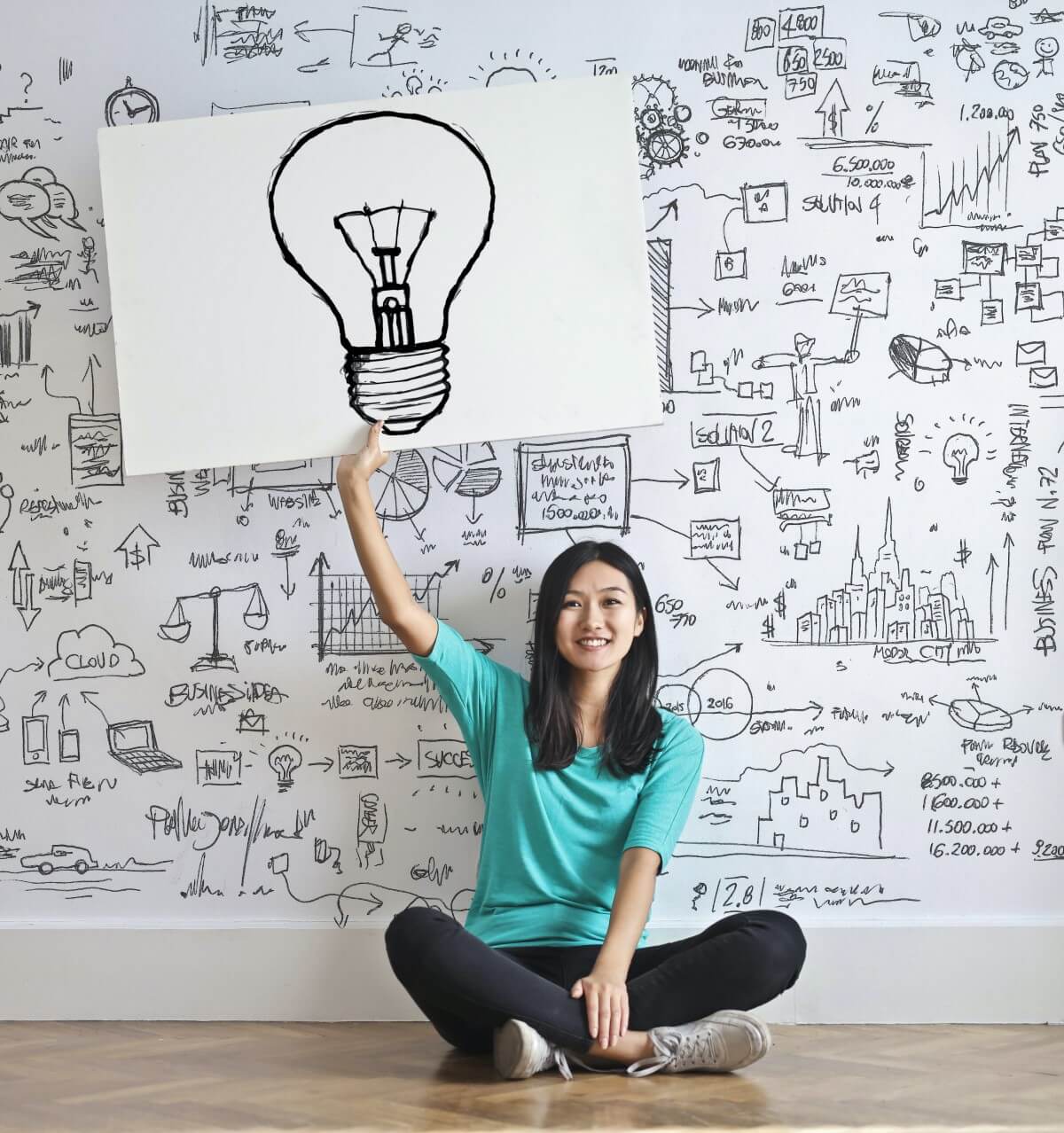 A portrait of a young Asian female sitting cross-legged on the floor against a white wall filled with equations and charts, holding up a placard with a light bulb scribbled on it.