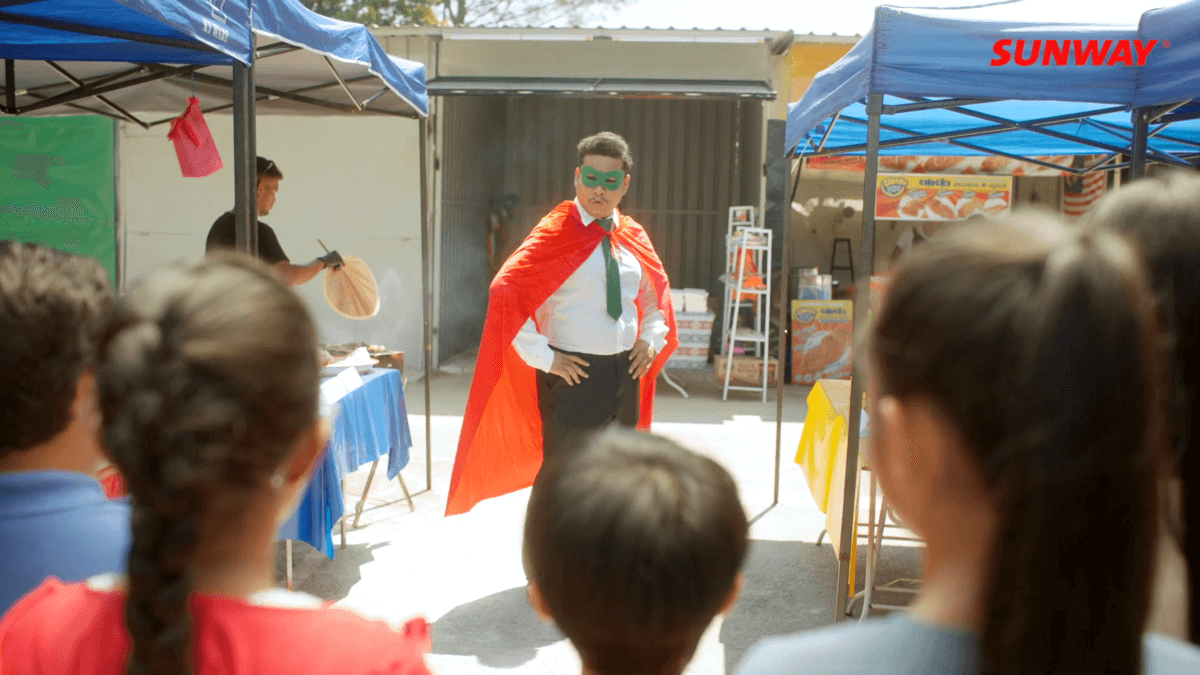 Abang Hero, clad in formal attire, green tie and mask and a red cape, with the 5 children in the foreground.