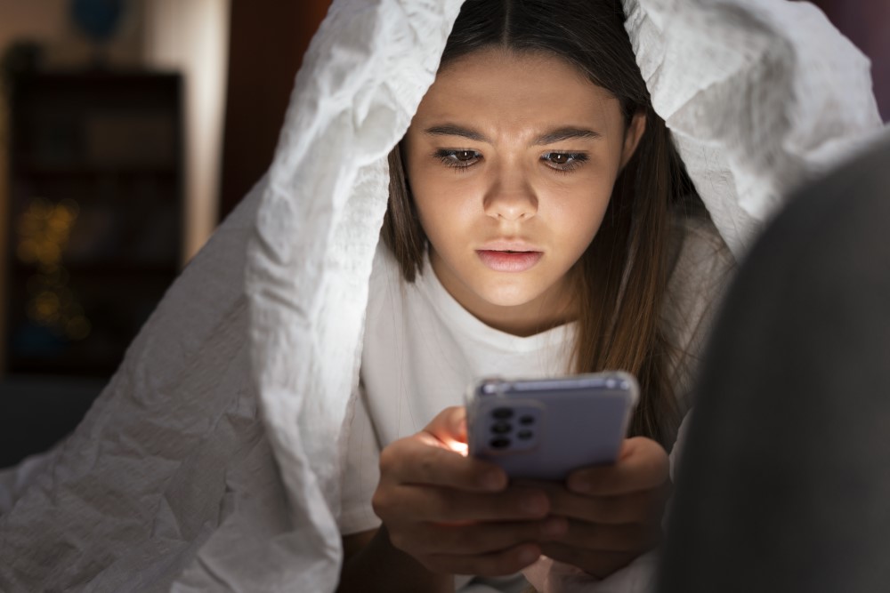 Medium shot suffering teenager being cyberbullied, under her blanket and holding her phone