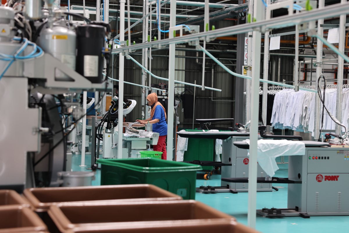 A full landscape shot of the Sunway Pristine Laundry plant garment section featuring a staff ironing clothing using a Sankosha industrial steam press as the focal point.