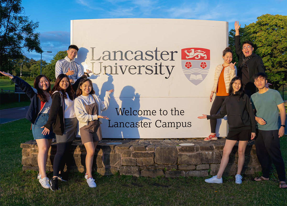 Evelyn Wee and some students posing in front of the Lancaster University signage, amidst greenery.