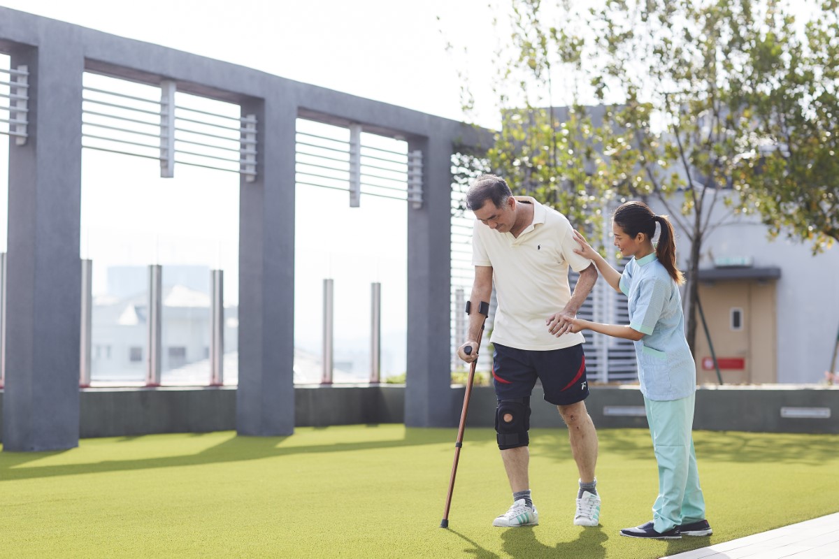 A nurse assisting a crutched elderly man in the gardens of Sunway Medical Centre, amidst greenery and a tree in the background.