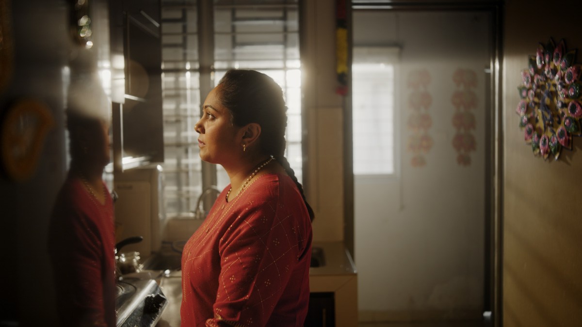 A mid-shot of an older lady looking solemnly out the window, with the morning sun in the background