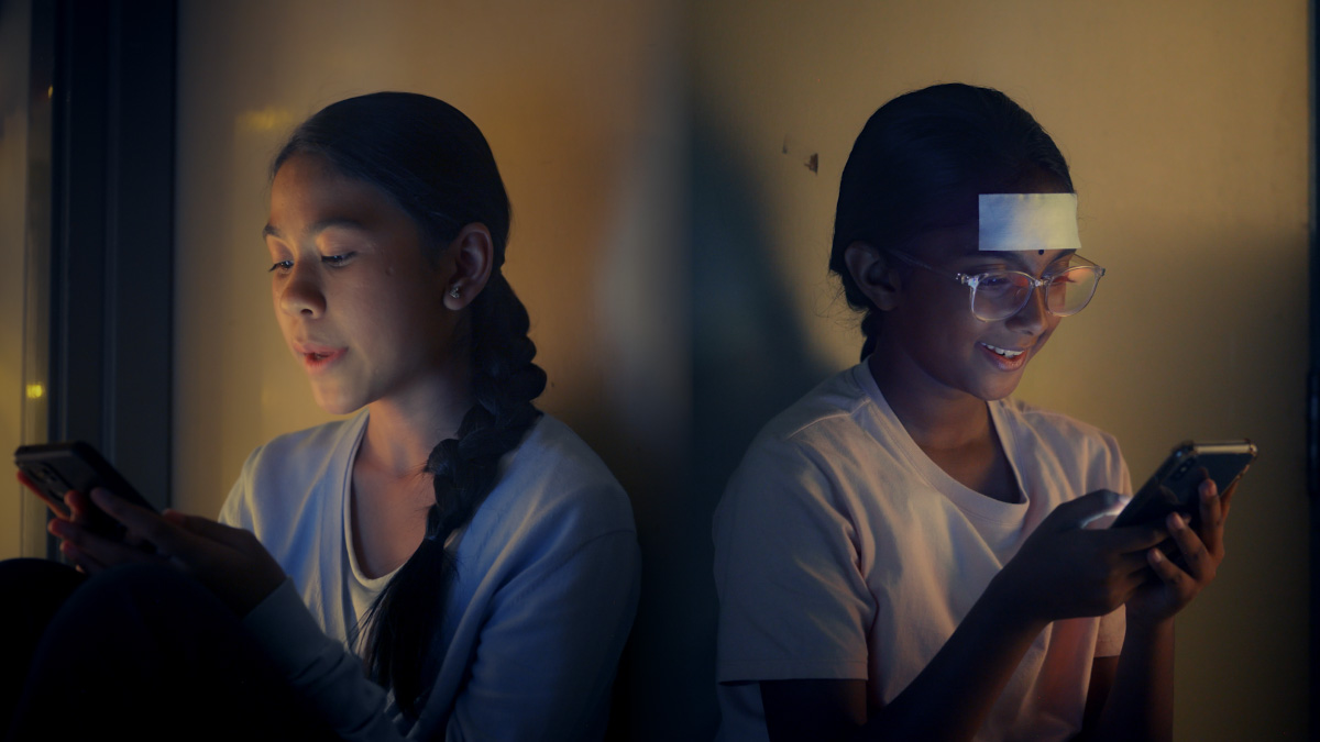 A mid-shot of two young ladies on their phones in a dimly lit room, smiling as they are texting